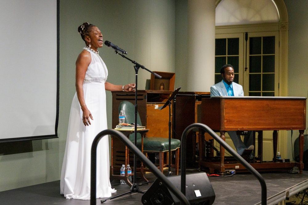 A woman in a white dress sings into a microphone on stage, while a man in a light blue shirt plays a keyboard in the background. They are performing in a room with green walls and a white column, and a screen is visible to the left.