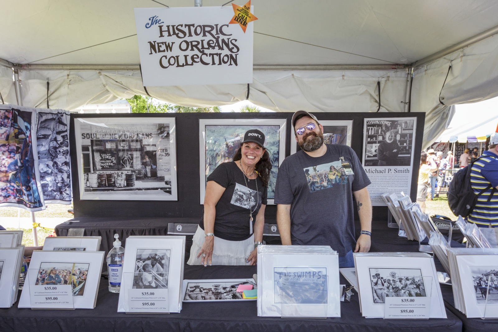 Two vendors stand under a tent displaying black and white photographs. A sign reads The Historic New Orleans Collection. The table showcases framed photos for sale, with prices visible. The vendors are smiling and wearing casual clothing.