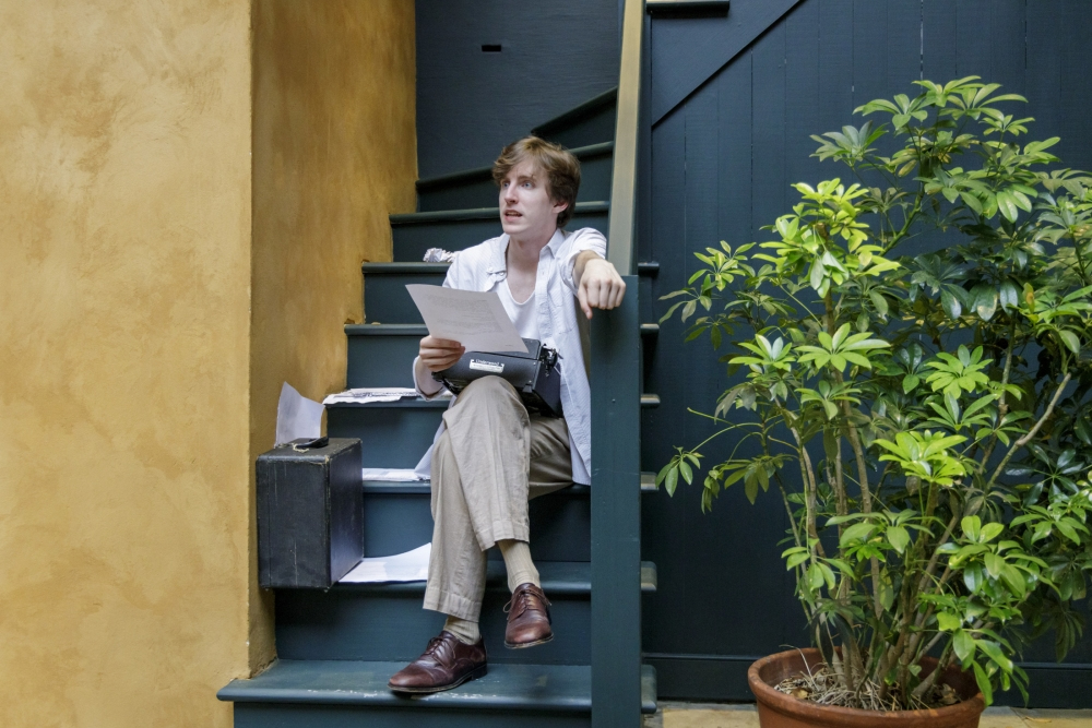 A person sits on green wooden stairs, holding papers and a typewriter. They wear a white shirt, beige pants, and brown shoes. Theres a potted plant nearby. The setting is domestic and casual.
