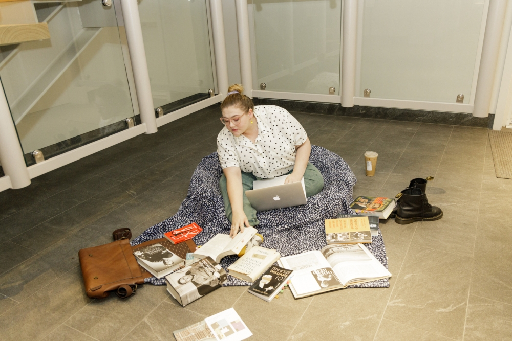A person sitting on a blanket in a library, surrounded by open books and a laptop. They are reaching for a book while a coffee cup and a pair of shoes are nearby.