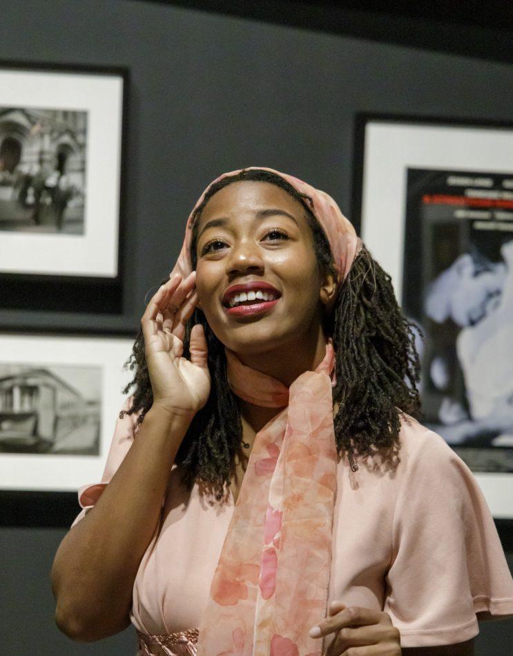 A woman with long braided hair and a pink scarf smiles while gesturing indoors. She is surrounded by framed black-and-white photos on a dark wall.