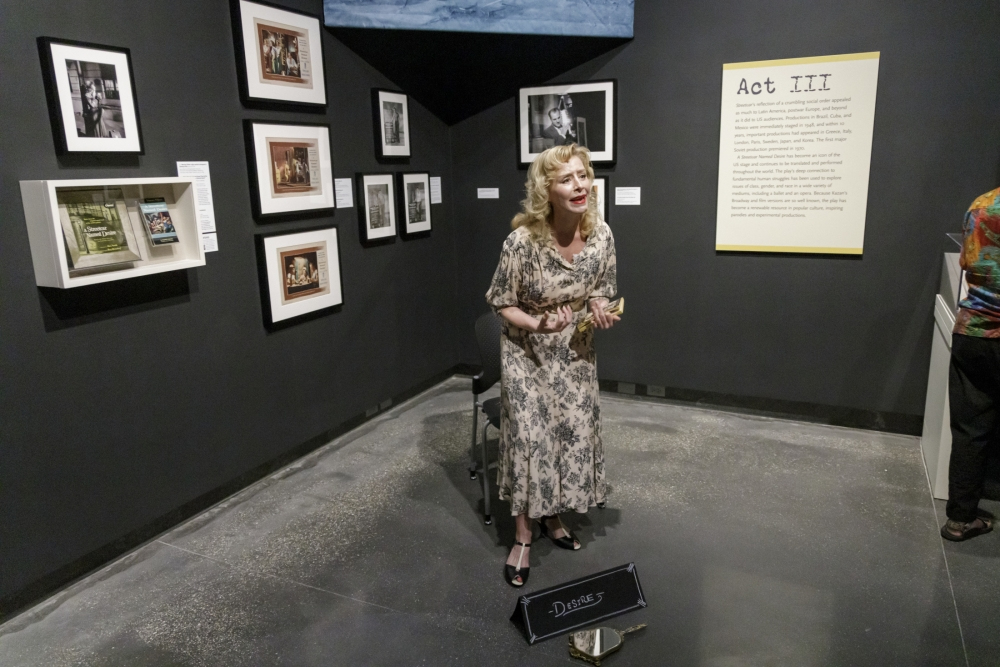A person with light-colored hair, wearing a floral dress, stands in an art gallery. They are smiling and holding an object while sitting on a stool. The wall behind them displays framed photographs and a sign titled Act III. A Donations box is on the floor.