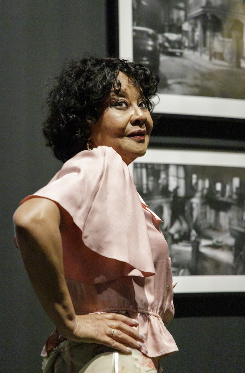 A woman with curly hair poses confidently, wearing a pink blouse. She stands in front of a black and white photograph on the wall, which is part of a dimly lit gallery or exhibition space.