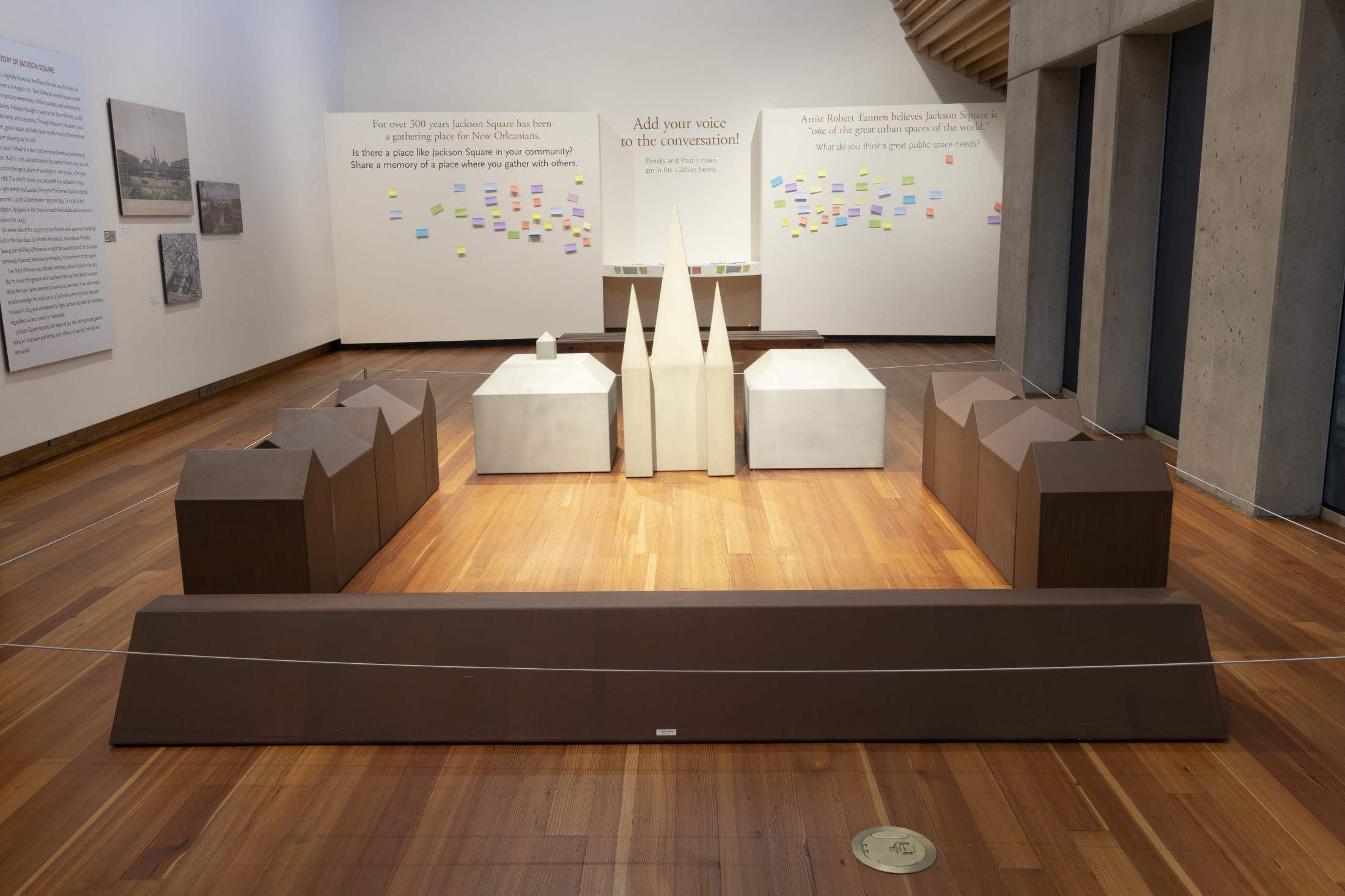 A minimalist museum exhibit displays a small-scale town model with gray geometric buildings on a wooden floor. Walls are adorned with informational posters and colorful sticky notes, inviting visitor interaction.