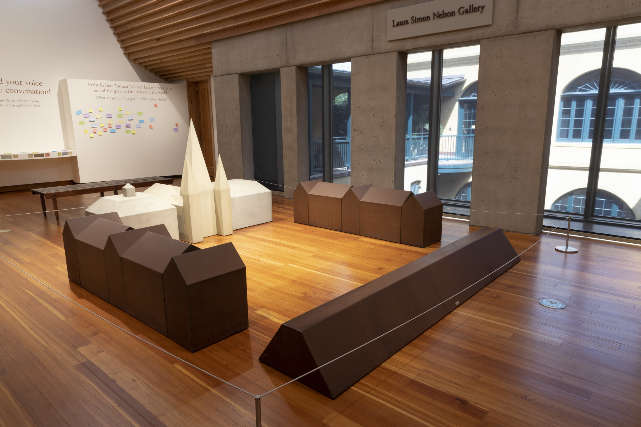 A gallery room with wooden floors features geometric sculptures resembling abstract buildings. The setup includes brown and white structures arranged in a semi-circle. Large windows allow natural light to filter in from the right.