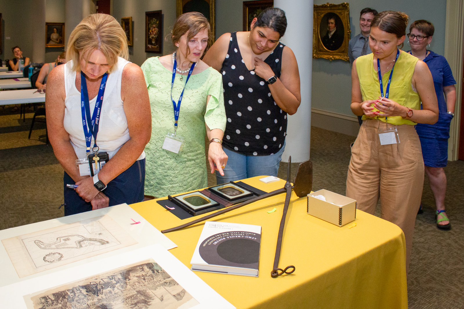 A group of people stand around a table covered with various artworks and objects in a gallery. One person points at a framed item while others observe closely. They wear name tags and seem engaged in a discussion. Paintings hang on the walls behind them.
