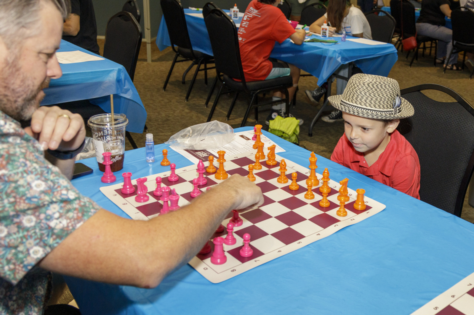 A man and a young boy play chess on a colorful pink and orange board at a tournament. The boy, wearing a straw hat, watches the mans move intently. Other participants are visible in the background at blue-covered tables.