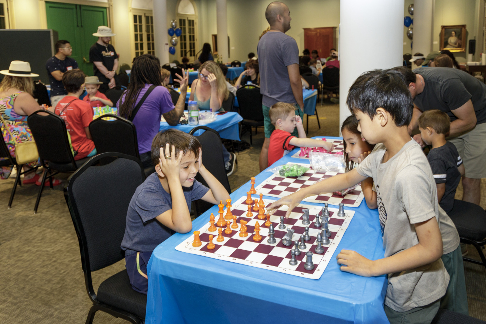 Children are playing chess at a table covered with a blue cloth in a busy room. One child looks thoughtful while contemplating a move. Adults and other children are engaged in activities at surrounding tables.