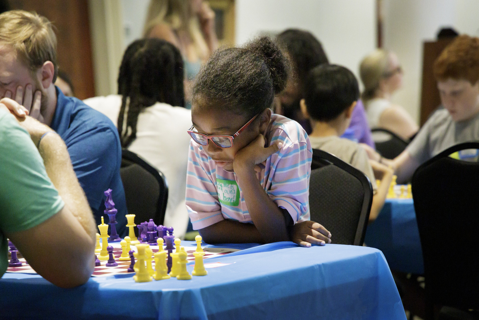 A young girl in glasses, wearing a striped shirt, thoughtfully playing chess with purple and yellow pieces. She is sitting at a blue table among other concentrated players in a room with several chess games in progress.