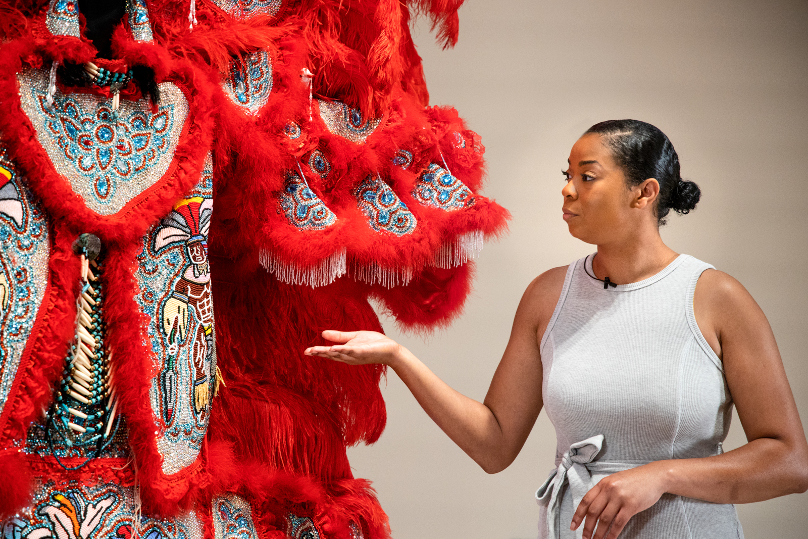 A woman in a gray dress gestures towards a vibrant red costume adorned with intricate beadwork and feathers, possibly part of an exhibit. The background is neutral, highlighting the detailed artistry of the costume.