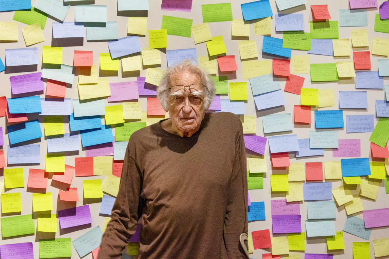 An older man with white hair stands in front of a wall covered with colorful sticky notes. He is wearing a brown long-sleeve shirt and leaning slightly. The notes vary in colors, including pink, blue, yellow, and green.