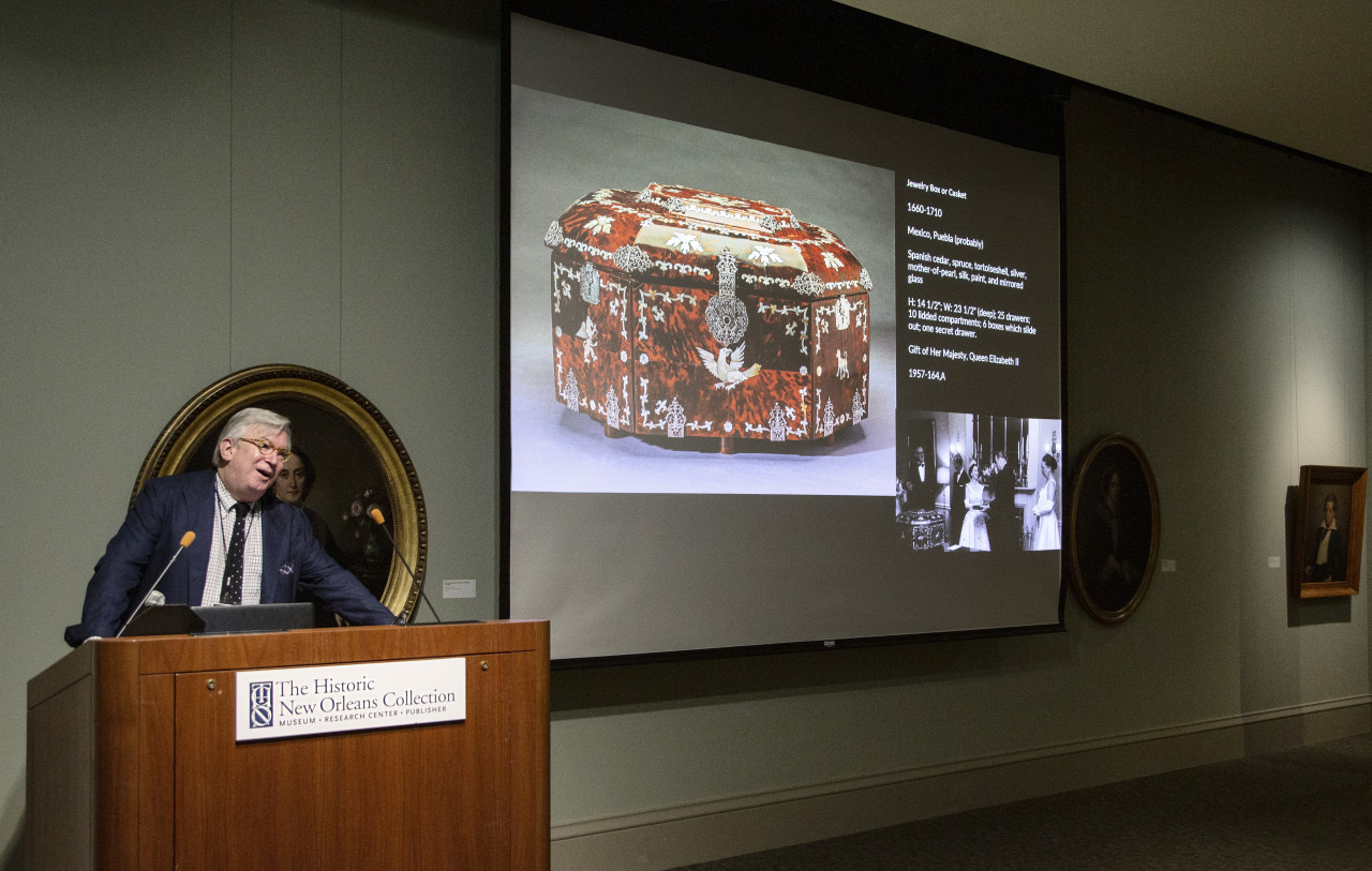 A speaker presents at a podium, displaying a slide of an ornate, decorated box. The slide includes text and a historical photo. The presentation is held at The Historic New Orleans Collection, as indicated on the podium. Portraits adorn the walls.