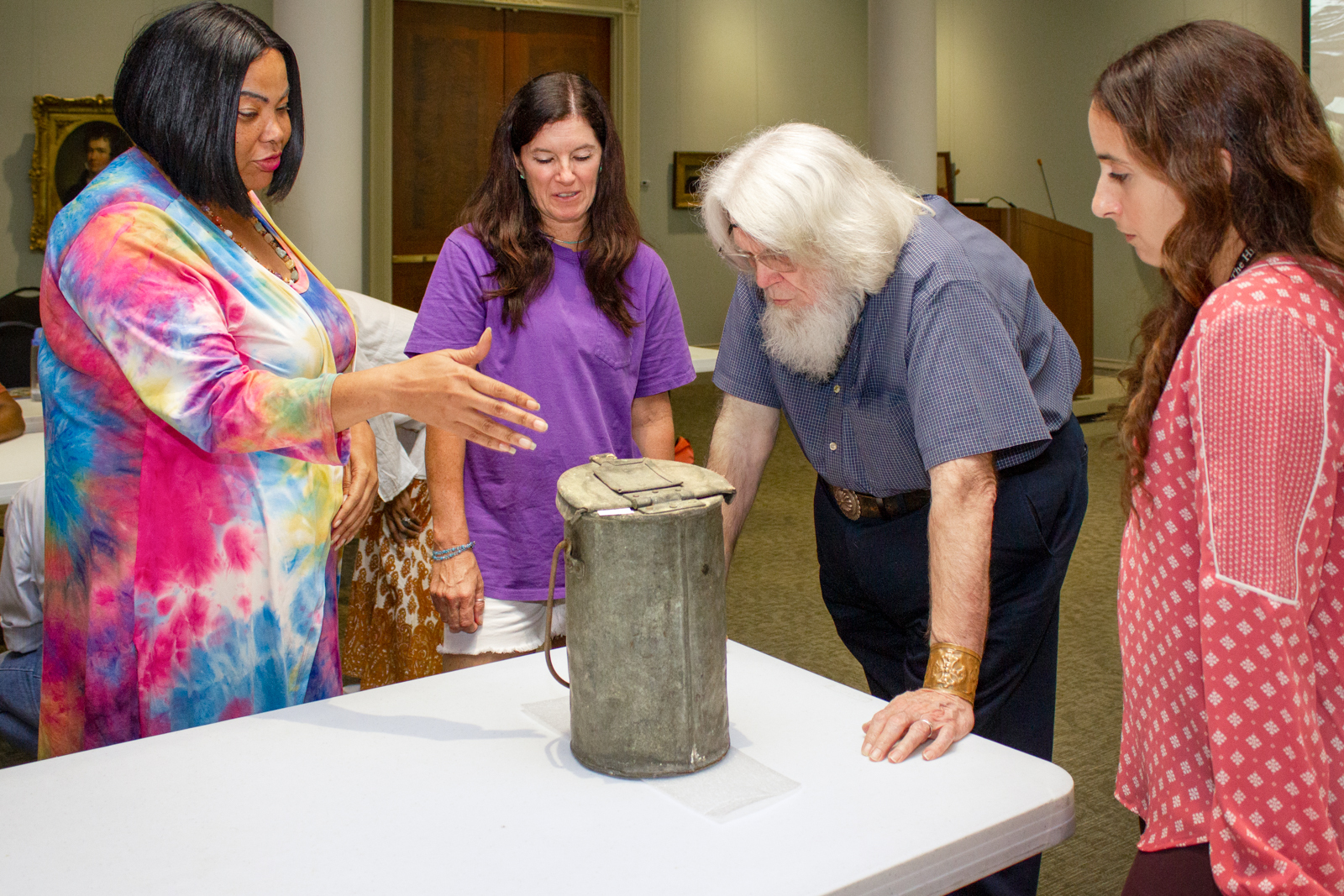 Four people stand around a table examining an old, weathered container. One person gestures towards it while the others observe closely. Theyre indoors with paintings on the wall.