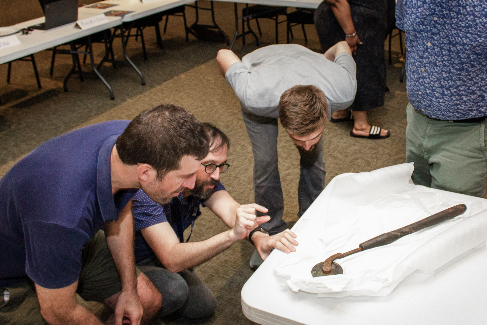 Three people are gathered around a table, closely examining a wrapped artifact, which looks like an old tool or weapon. They appear to be in a meeting room, with other tables and chairs in the background.