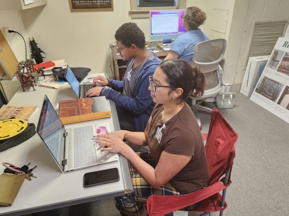 Three people working on laptops in a small room with books and posters around. Two are seated at a table, and one is in a folding chair. The room has a cozy and studious atmosphere.