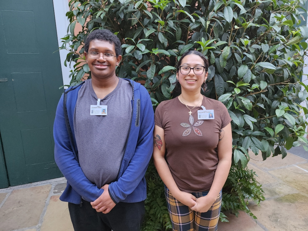 Two people smiling and standing side by side in front of a large leafy plant. The person on the left wears glasses and a blue hoodie, while the person on the right wears glasses and a brown shirt with a leaf design. Both have ID badges.