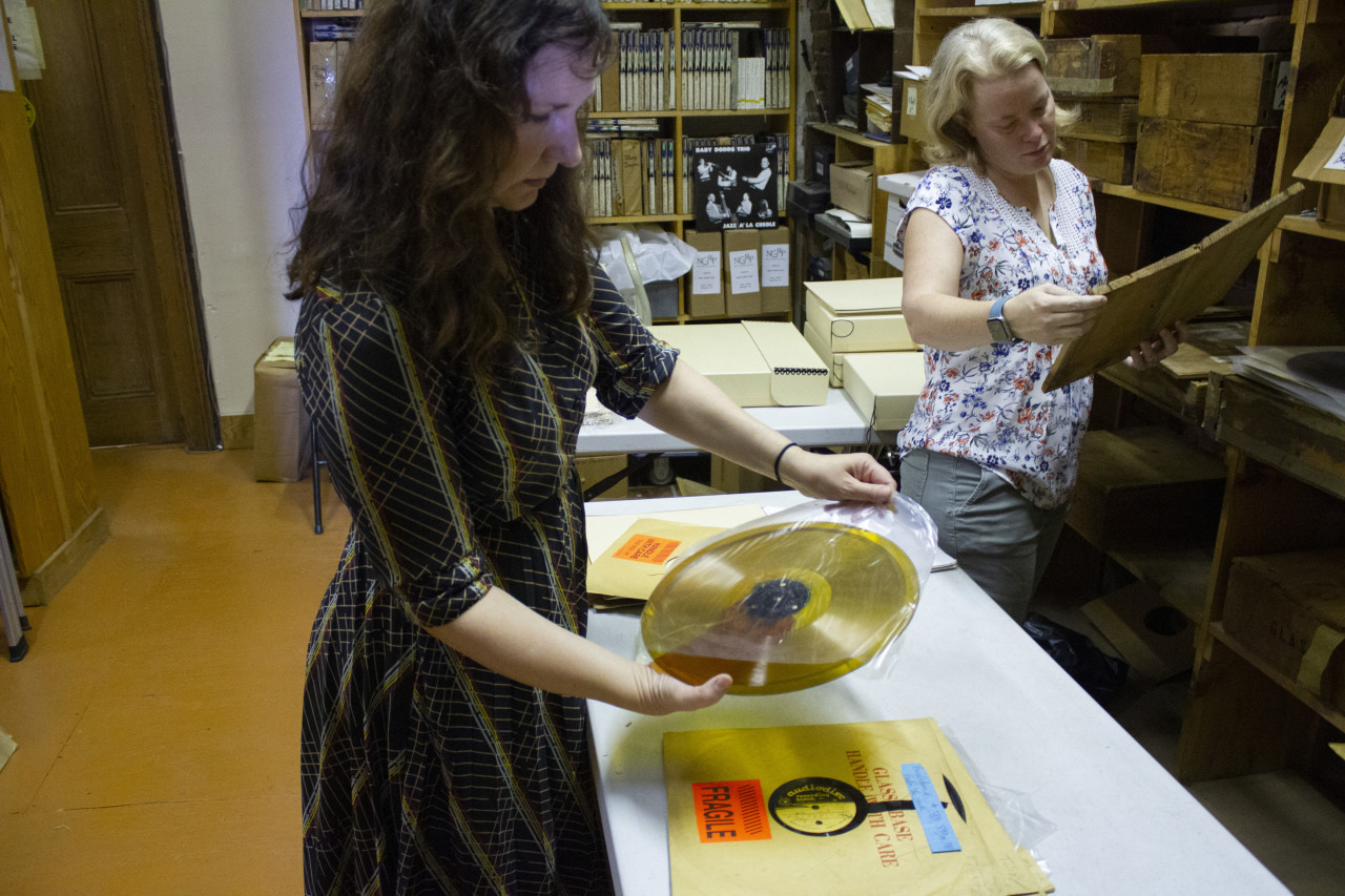 Two women in a storage room examine vinyl records. One woman holds a record with a label; the other checks records in an envelope. Shelves with boxes and records surround them, creating an archive-like setting.