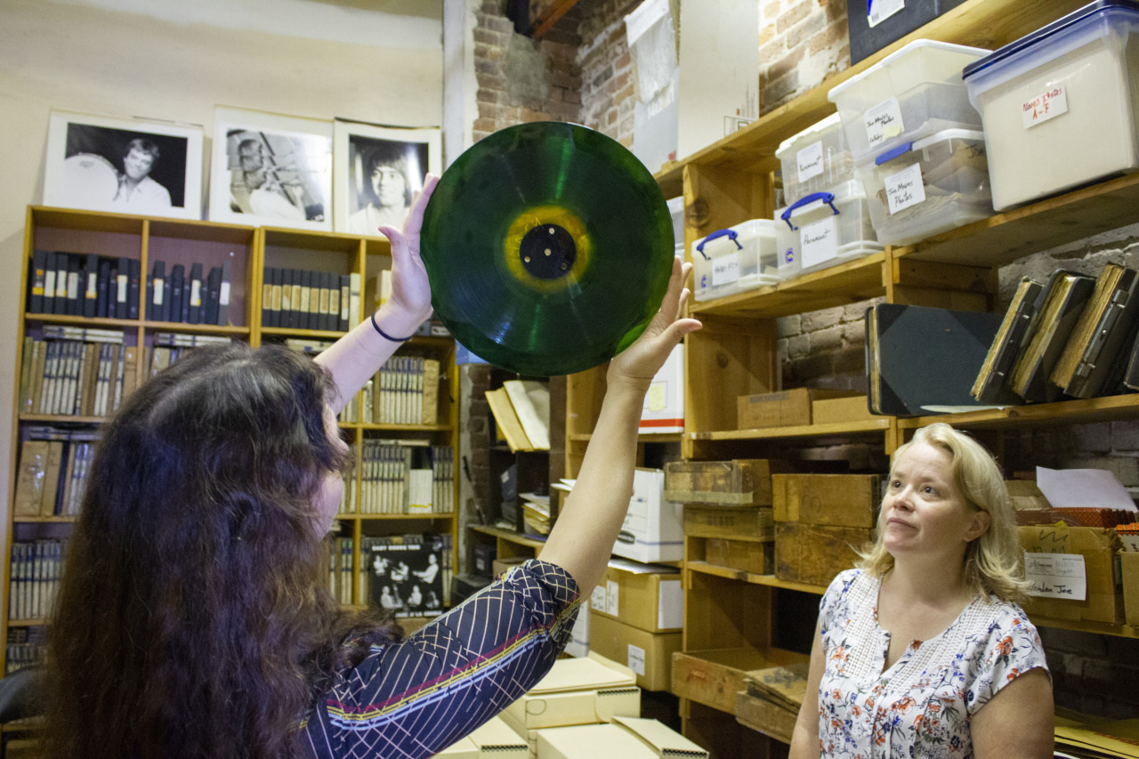 Two women in a room full of shelves with records and boxes. One woman holds up a translucent green vinyl record, admiring it. The other woman looks on. Black-and-white photos are on the wall, and the shelves are stacked with various items.