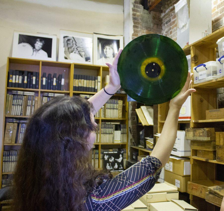 A woman with long hair holds up a translucent green vinyl record in a room filled with shelves of vinyls, boxes, and framed black-and-white photos.
