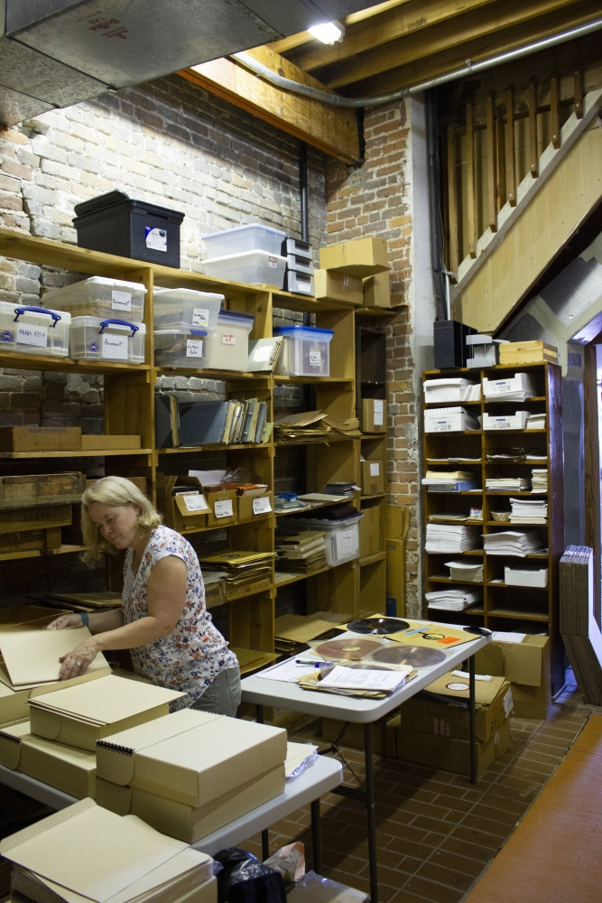 A person is organizing archival materials in a storeroom filled with shelves of documents and boxes. The space has brick walls and a wooden staircase. The person is handling folders on a table, surrounded by various archival supplies.