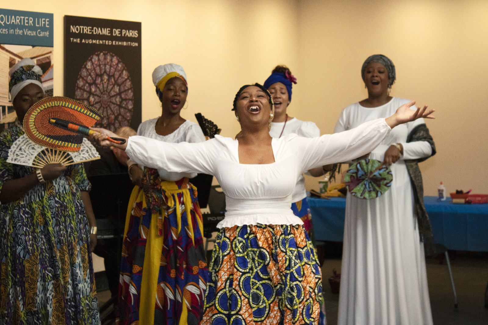 A group of women passionately singing and dancing in colorful traditional attire. One woman, in the center, has her arms spread wide, holding a fan. Background features a Notre-Dame de Paris exhibition poster.
