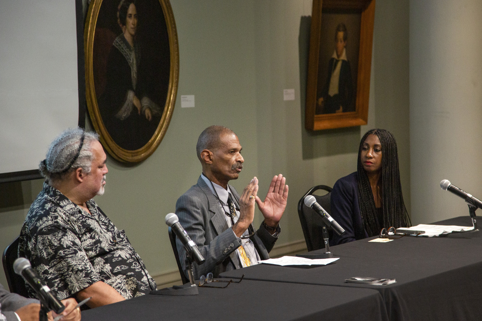 Three people seated at a table participate in a panel discussion. A man in the middle gestures with his hands while speaking. The other two listen attentively. Portraits hang on the wall behind them, and microphones are on the table.