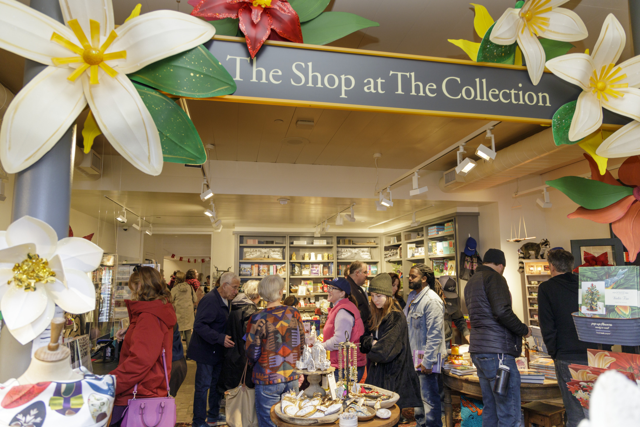 A busy gift shop with customers browsing various items. The entrance is decorated with large, colorful flower sculptures and a sign reading The Shop at The Collection. Shelves display books, gifts, and decorative items.