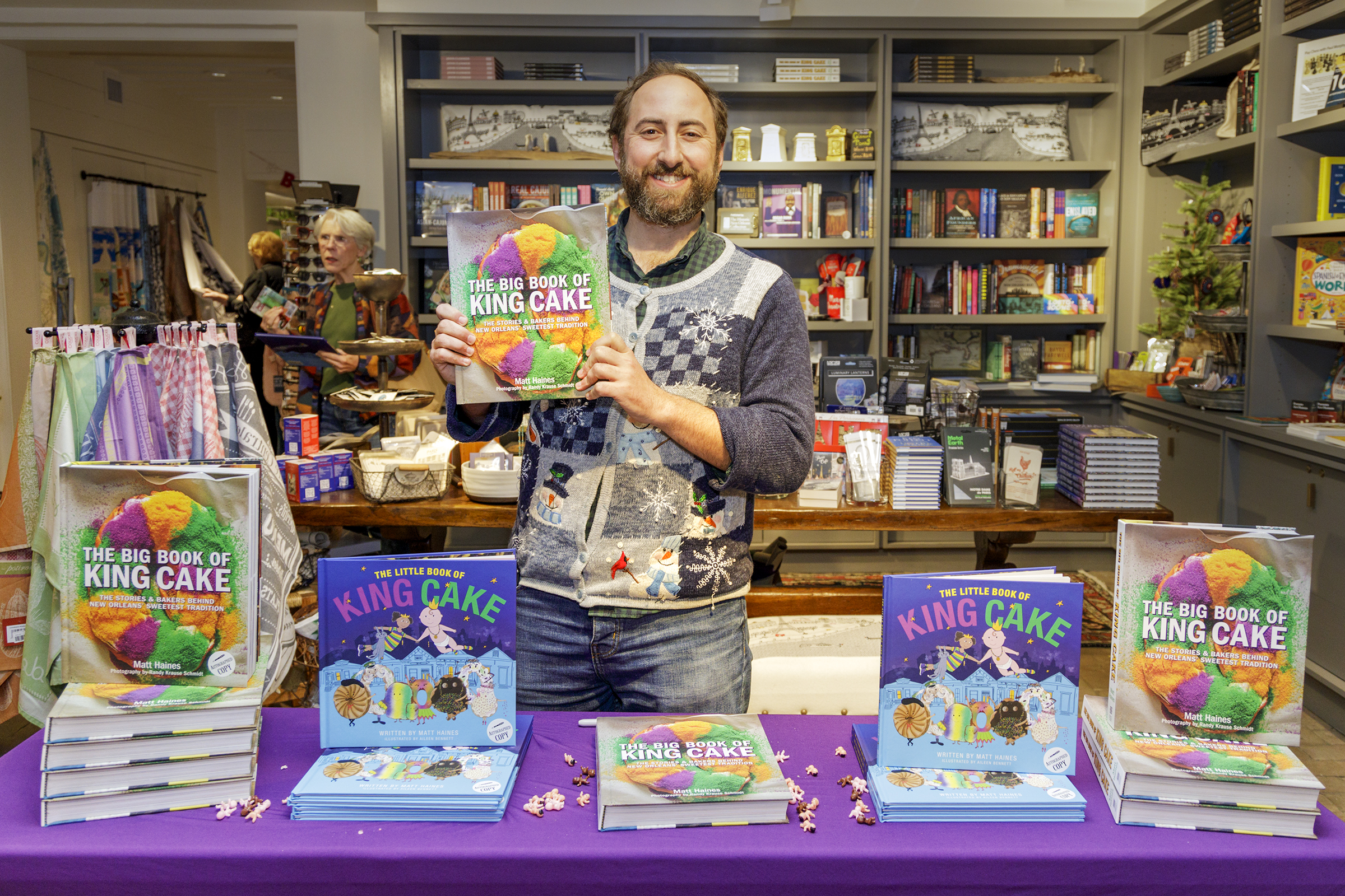 Author Matt Haines smiles and holds up his Big Book of King Cake while standing behind a table in the Shop at the Historic New Orleans Collection.