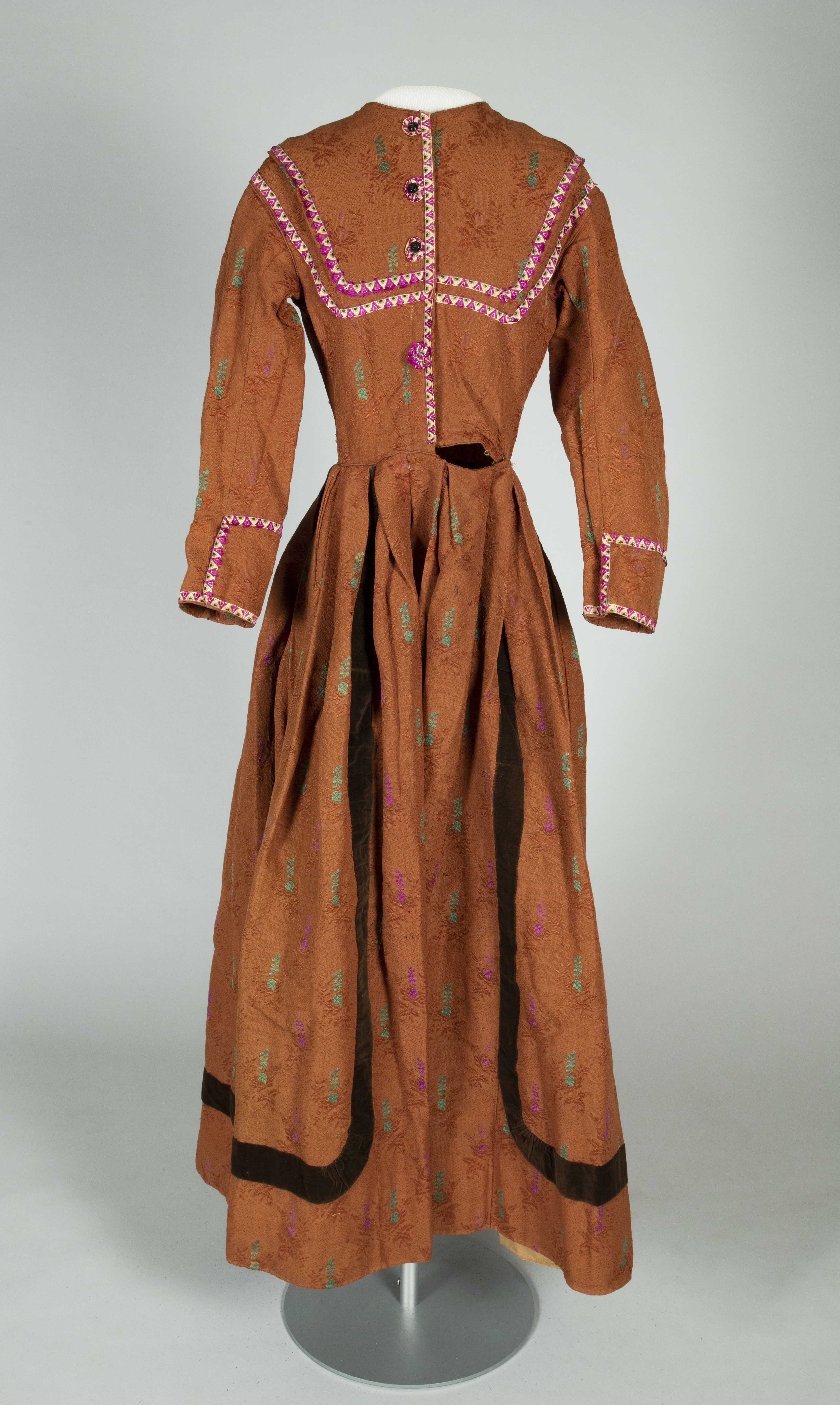 A vintage brown dress on a mannequin, featuring a full-length skirt, long sleeves, and intricate embroidery with pink trim on the neckline and cuffs. The dress has a textured fabric with floral patterns and a dark band near the hem.