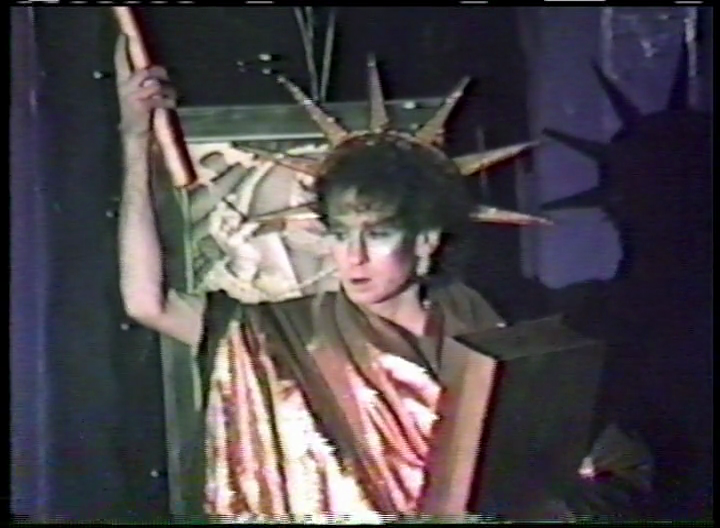 A person dressed as the Statue of Liberty holds a torch and book against a dark background, with a statue or painting partially visible behind them. The outfit includes a spiked crown and metallic draped clothing.