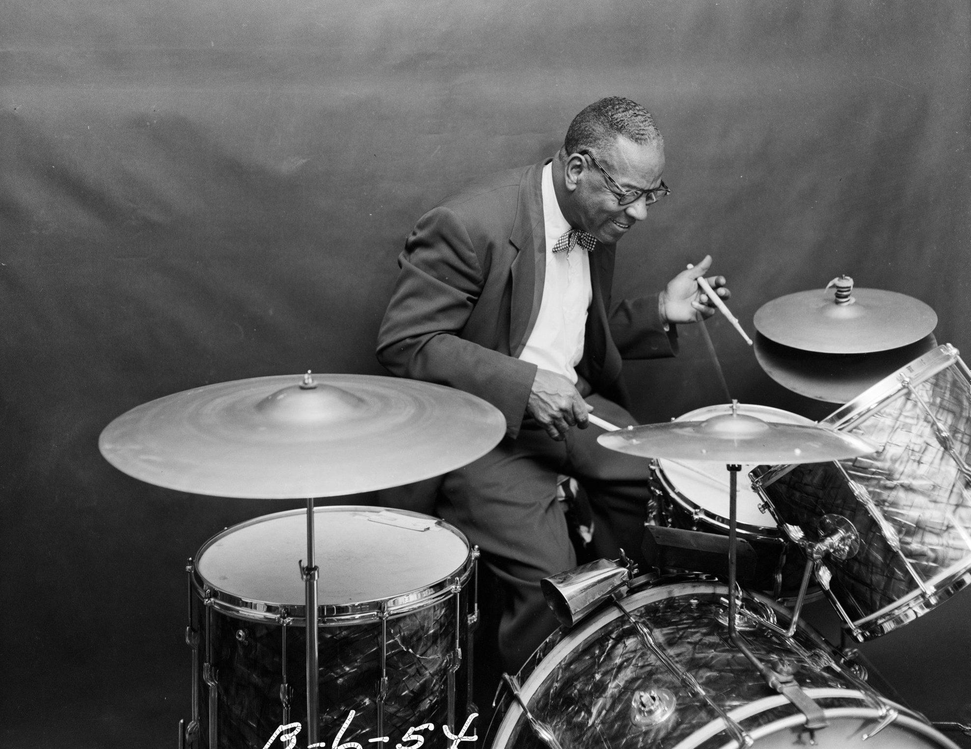 A man in a suit and bowtie is playing a drum set with a focused expression. The background is plain and the image is in black and white.