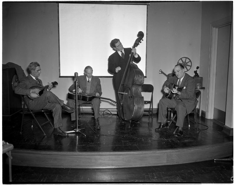 Four men are seated on a small stage, playing musical instruments. One plays the mandolin, another the slide guitar, a third stands and plays the double bass, while the fourth plays the guitar. A microphone is positioned in front of them.