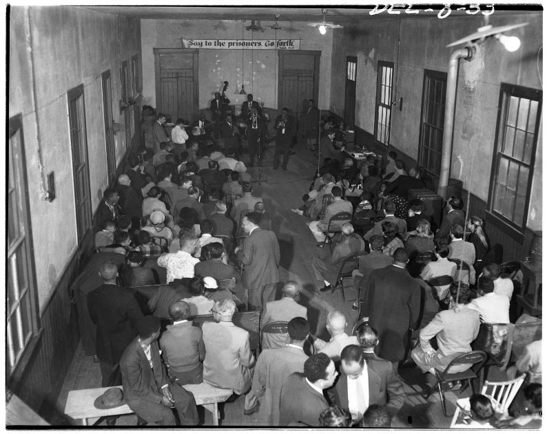 A black and white photo of a crowded room with people seated and standing, facing a stage where musicians are performing. A sign above the stage reads, Sing to the prisoners, go forth. The room has high windows and wooden floors.