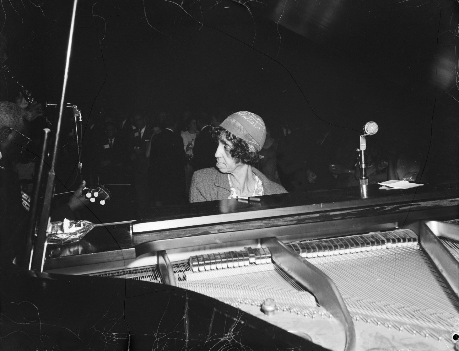 A close up black and white photograph of "Sweet" Emma Barrett playing the piano during a performance at an unidentified venue. She wears an embroidered skull cap that reads "Emma Bell Gal".