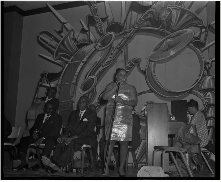 A woman in a shiny dress sings into a microphone on a stage. She is accompanied by musicians in suits sitting with instruments. The backdrop features a large mural of musical instruments.