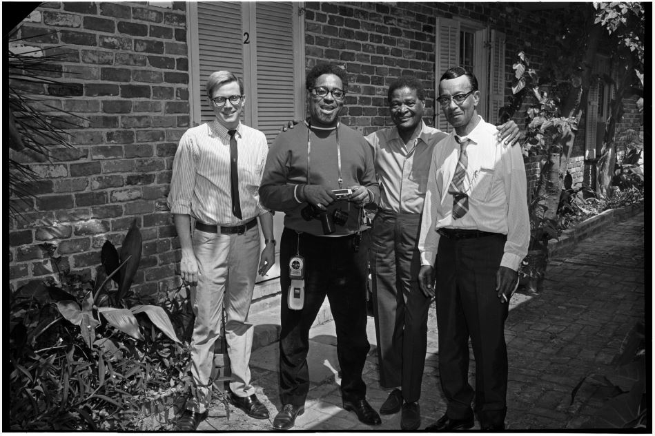Four men stand together outside in front of a brick wall. Three are wearing glasses. They are dressed in casual, professional attire and are smiling at the camera. One man holds a camera around his neck. Vegetation is visible nearby.