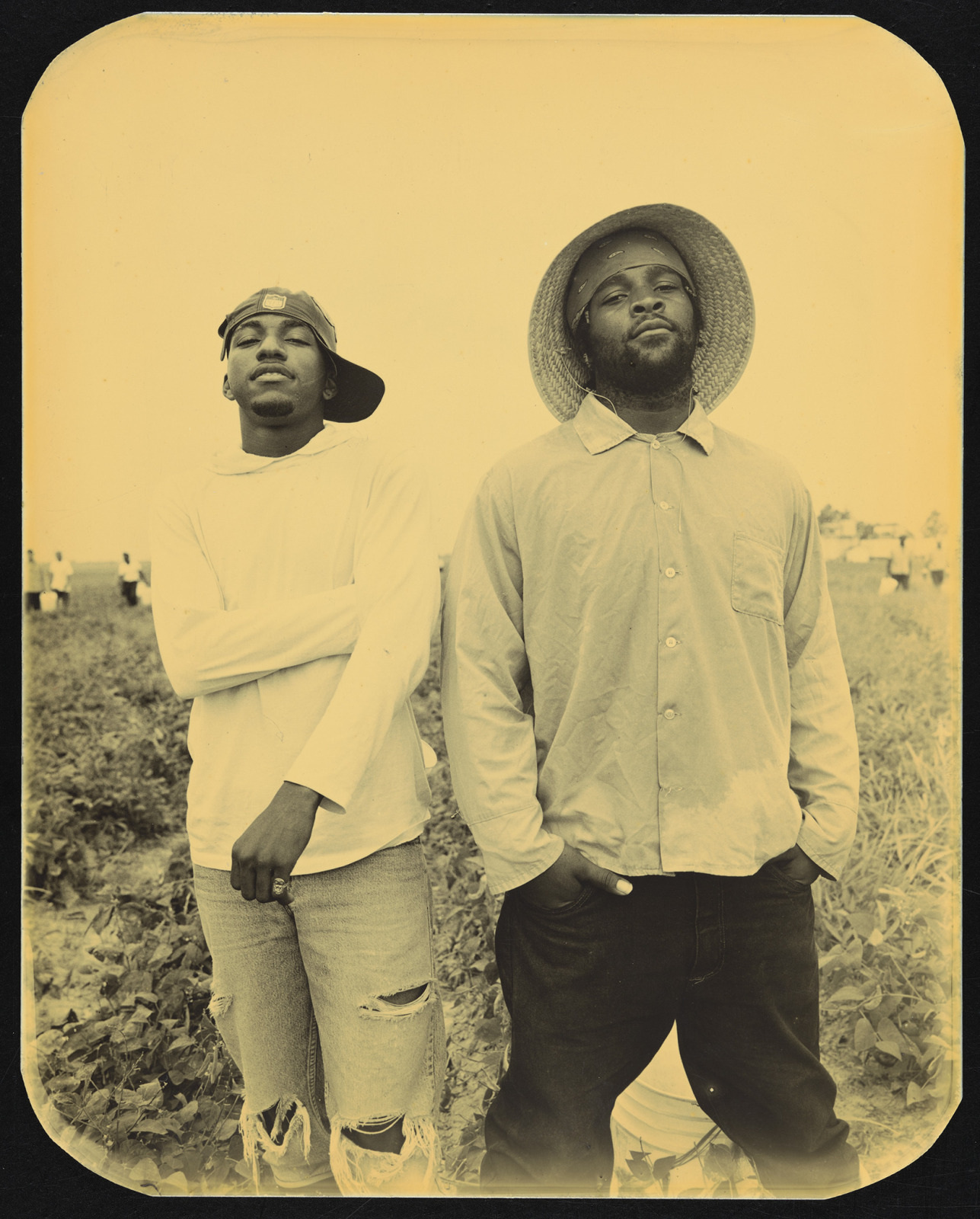 Two people stand confidently in a field. One wears a sideways cap and a torn shirt, arms crossed. The other wears a wide-brimmed hat and a buttoned shirt, hands in pockets. The image has a yellowish tint, giving it a vintage feel.