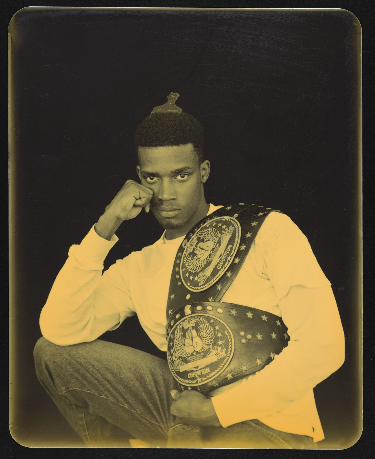 A sepia tone photograph shows an incarcerated young black man who is wearing a white long sleeve shirt and cradling two wrestling trophy belts. He is looking expressionless at the camera, with this head resting on his fist, which is resting on his right knee.