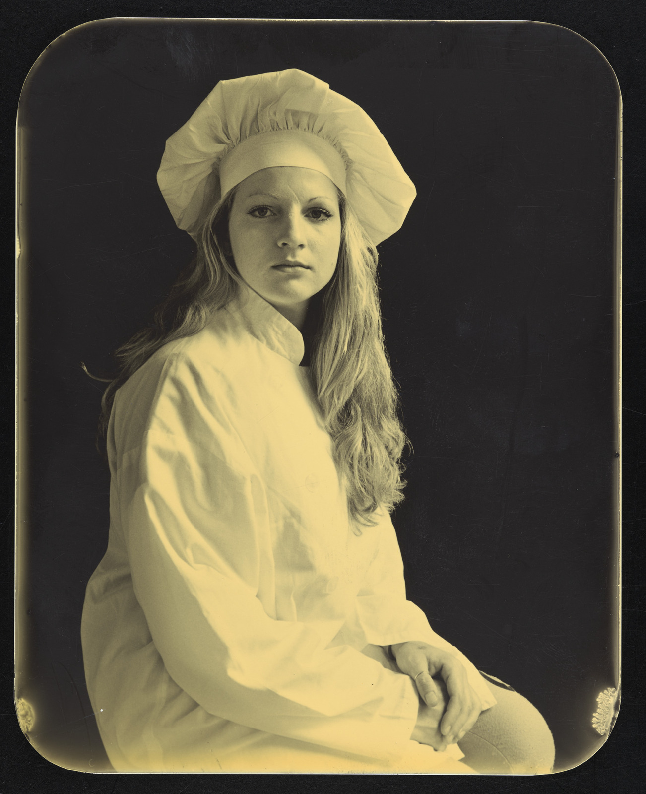 A sepia tone photograph shows an incarcerated, young white woman with blonde hair, who is wearing a white gown and wearing a chef's hat.