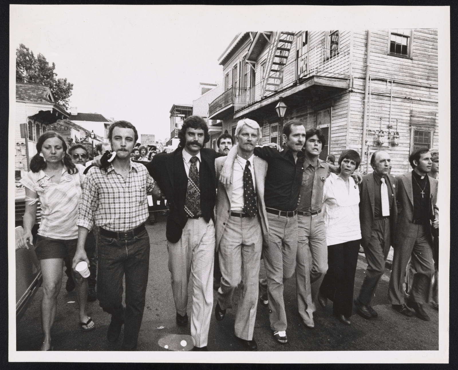 HERE demonstrators marching through the French Quarter in protest of Anita Bryant’s appearance at New Orleans’s Municipal Auditorium. A group of men and women march down the street, locking arms in solidarity, with French Quarter buildings visible in the background.