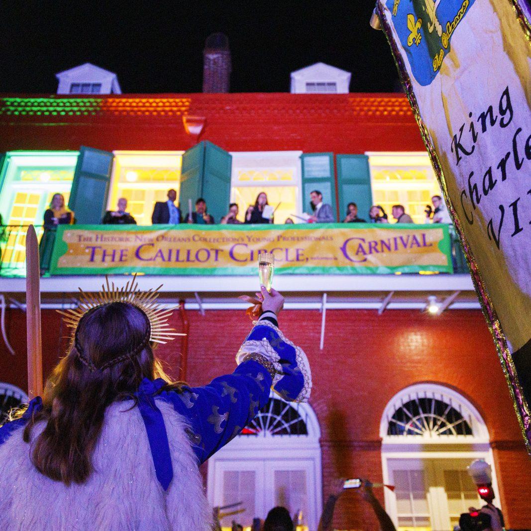 A person in festive attire raises a glass towards a balcony with a colorful banner reading Carnival at night. The crowd below watches, and the building is illuminated with vibrant lights. A banner on the right includes King Charles VI.