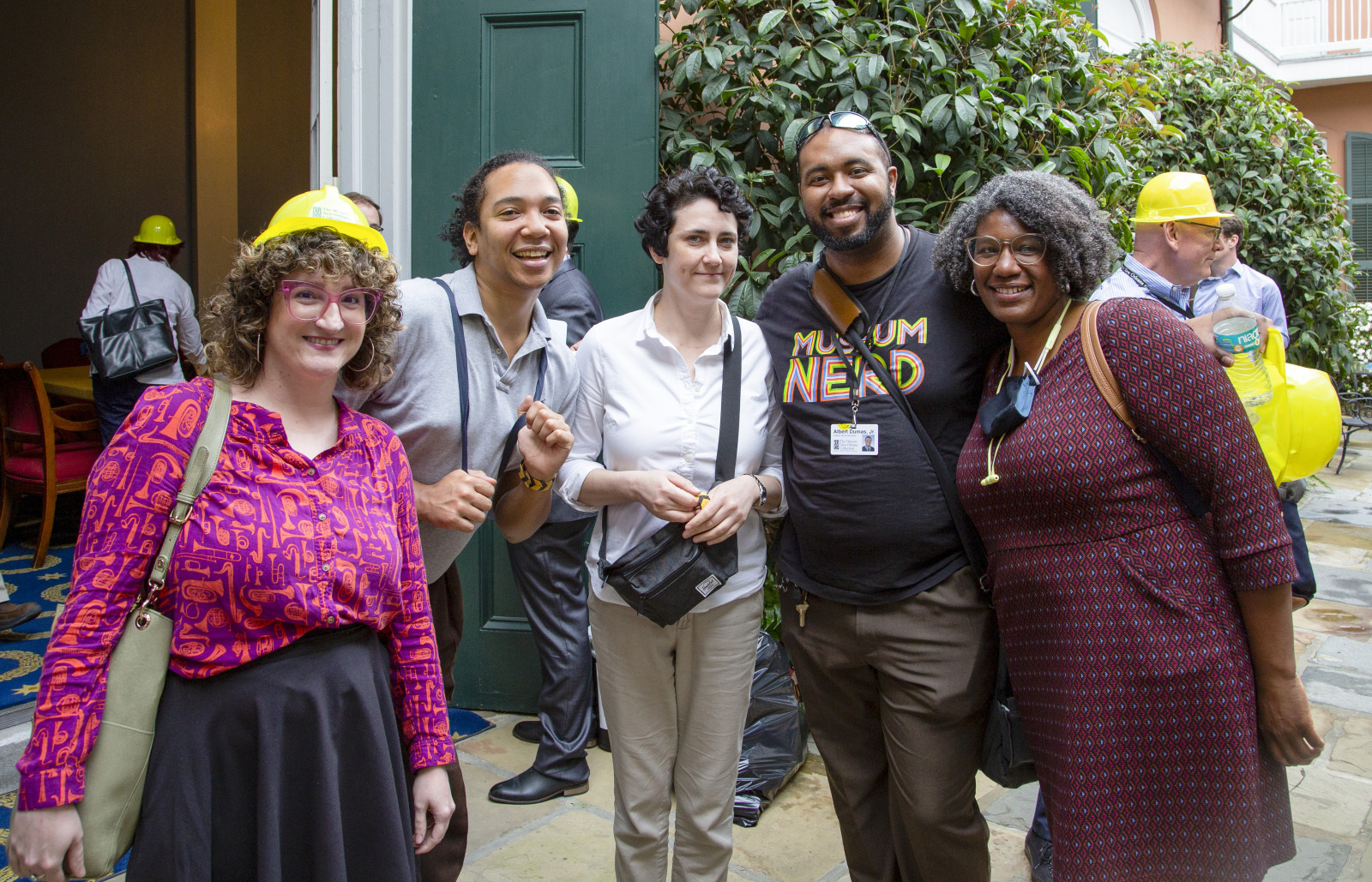 A group of six people smiling together outdoors. Some are wearing yellow hard hats, and one holds a walkie-talkie. They are standing in front of some greenery and an open door.