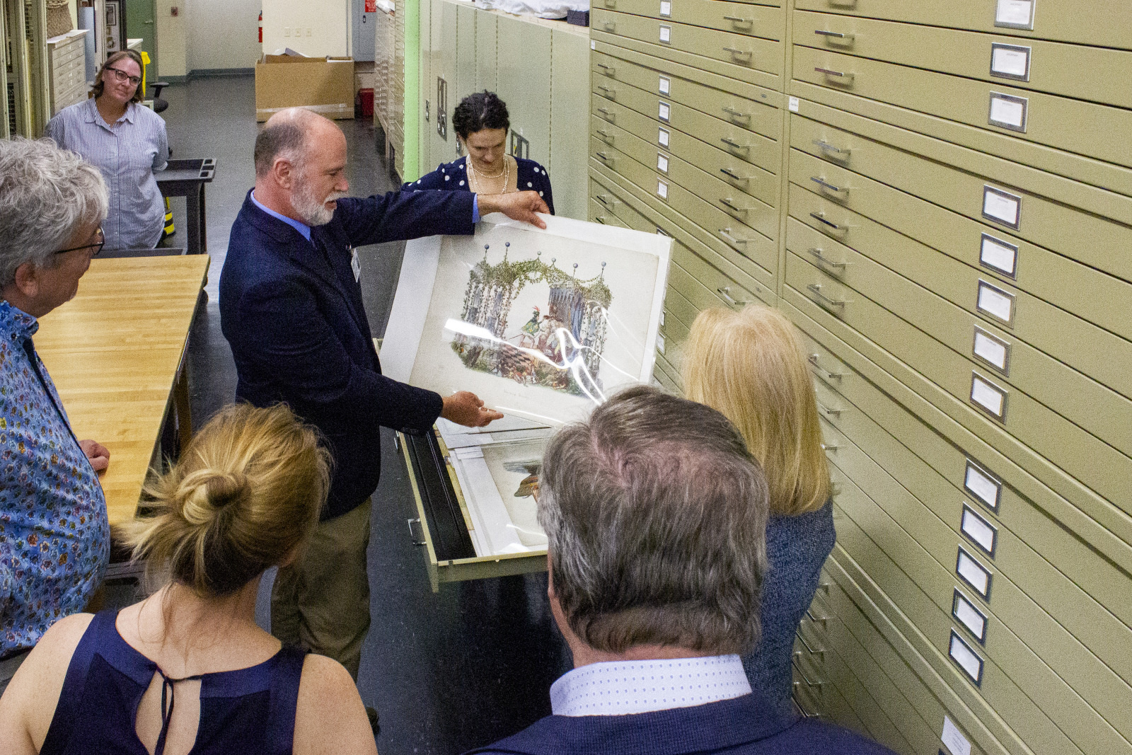 A group of people gather around a man holding an artwork in a storage room with large, labeled cabinets. The man shows the piece to the others, who watch attentively. Shelves and a wooden table are visible in the background.