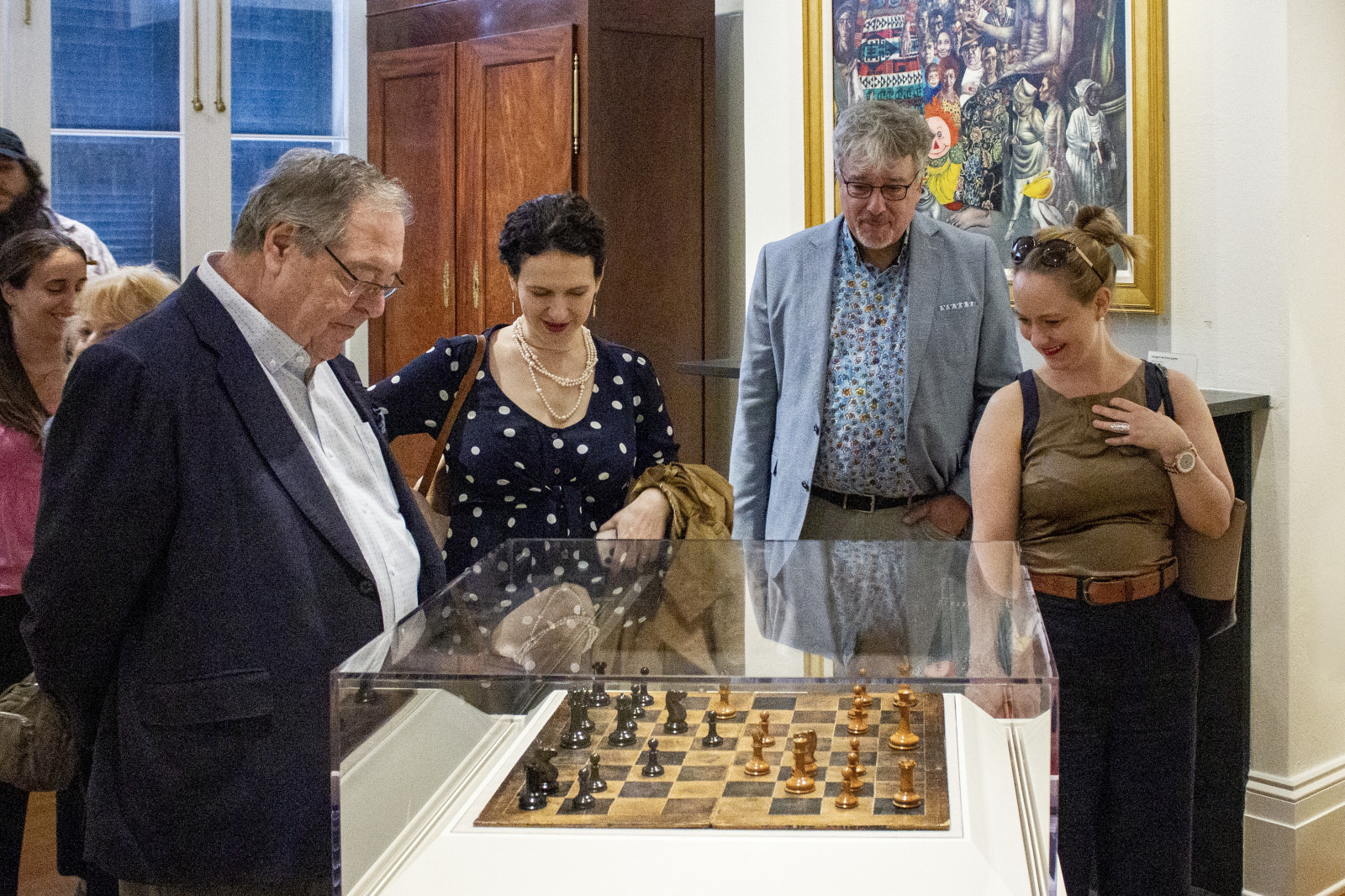 A group of people observes a glass-protected chessboard in a gallery. One man looks intently at the board, while others chat and smile. Various artworks are visible on the walls.