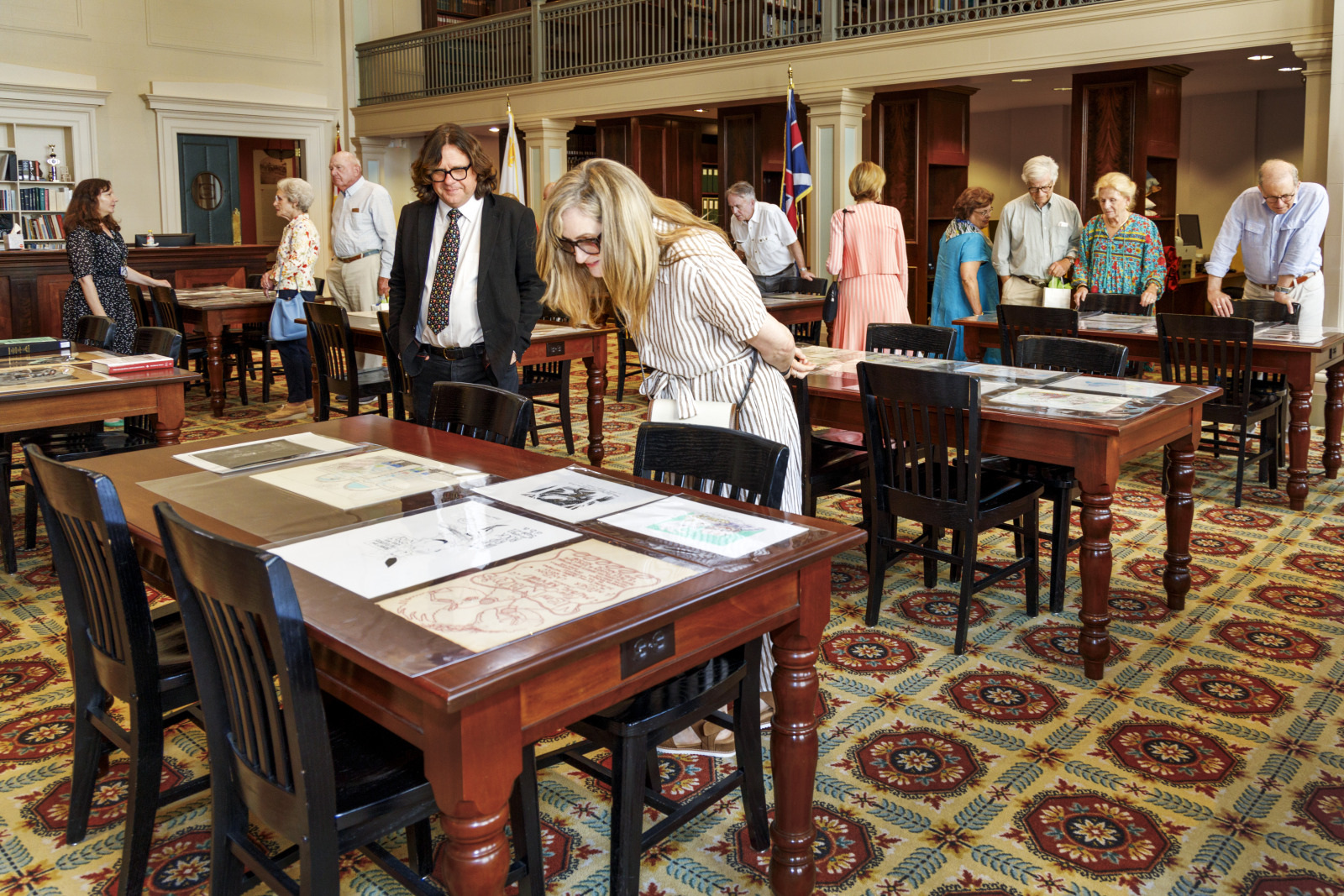 People are gathered in a library or museum setting, examining documents on wooden tables. The room has high ceilings, bookshelves, and a decorative carpet. Some individuals appear to be in conversation while others focus on the documents.