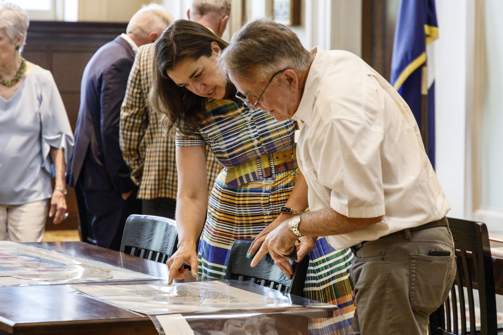 A woman and a man are examining maps on a table in a room with several other people. The woman is wearing a colorful dress, and the man is dressed in a light shirt and pants. An American flag stands in the background.