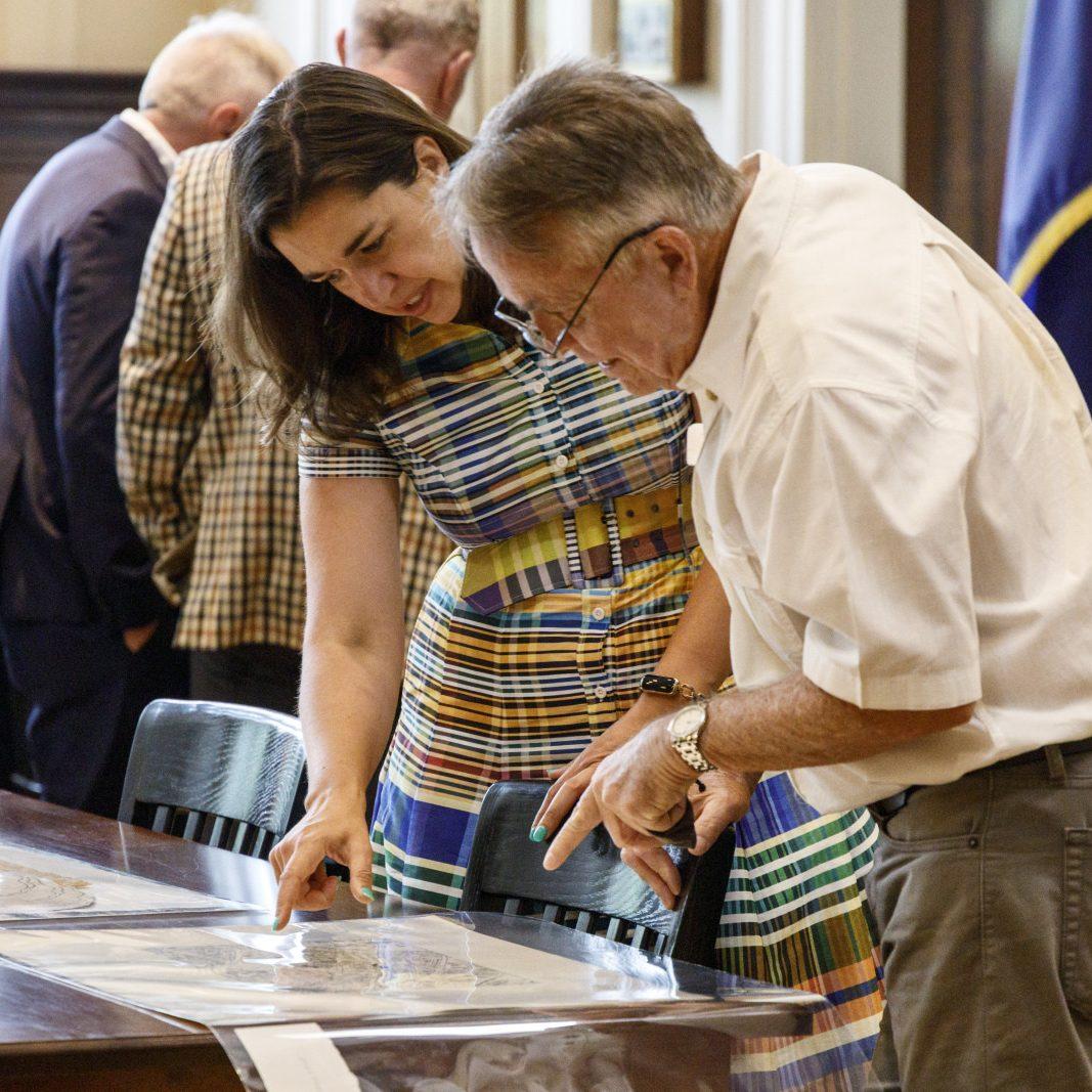 A woman in a colorful striped dress and a man in a white shirt examine a document on a table. They appear to be in a formal setting, with other people in the background.