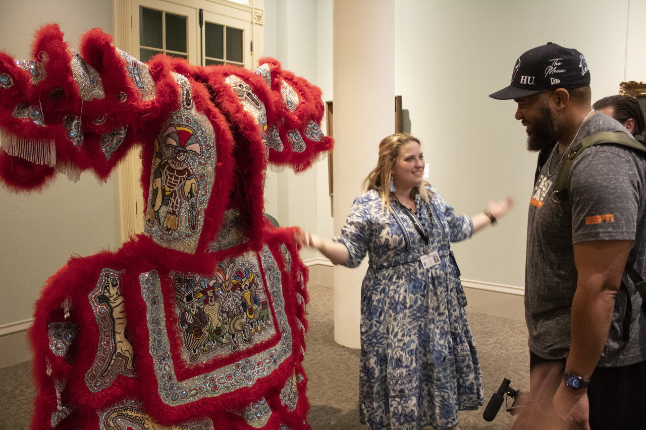 A smiling woman in a blue patterned dress gestures animatedly towards a standing man in a cap. Between them is an ornate red costume adorned with intricate beadwork and feathers. They appear to be inside a museum or gallery.