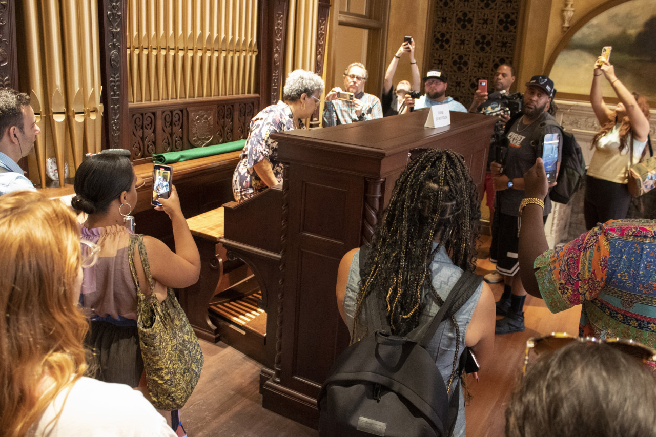 A group of people gather around a vintage organ inside a historic room. Many are taking photos with their phones. A person stands at the organ, engaging with the crowd. The room features intricate woodwork and an ornate organ facade.