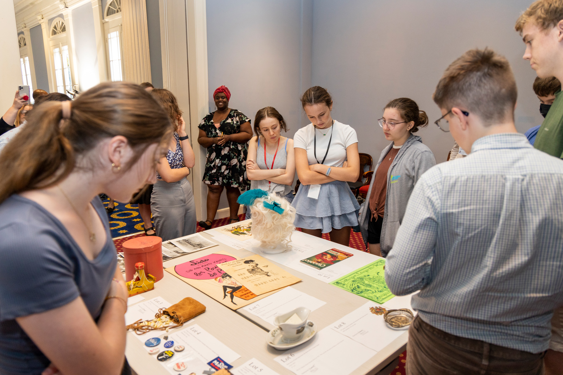 A group of teenage students gather around a table displaying various historic items, including posters, buttons, and a feathered mask. The setting appears to be indoors with ornate architecture in the background. People are observing and discussing the items.